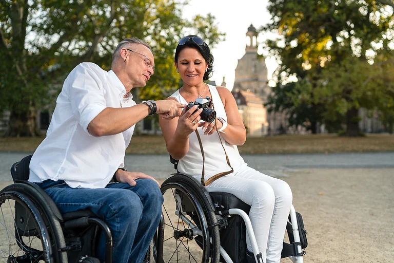 Zwei Rollstuhlfahrende schauen sich im Park ein Foto auf einer Kamera an, im Hintergrund unscharf die Frauenkirche