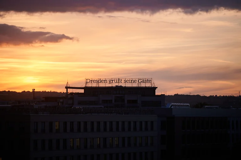 „Dresden grüßt seine Gäste“ steht als Schriftzug auf einem Dach, dahinter geht die Sonne unter und taucht die Szene in warmes Licht.