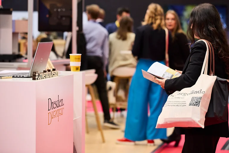 IMEX Frankfurt 2024, Seitenperspektive: Eine Messebesucherin steht mit Stoffbeuteln am Counter, vertieft in eine Broschüre. Links im Bild der Dresden Convention Counter mit Dresden-Logo, Holzschriftzug #DresdenConvention und einem Laptop. Im Hintergrund unscharf weitere Besucher:innen.