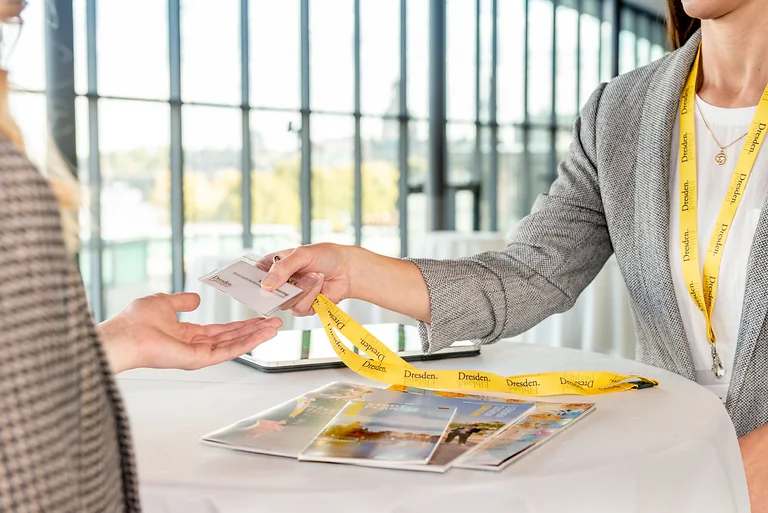 Übergabe eines Teilnehmer-Badges mit gelbem Dresden-Lanyard, auf dem Tisch liegen Broschüren. Im Bildausschnitt sind nur Hände und Oberkörper der Personen zu sehen.