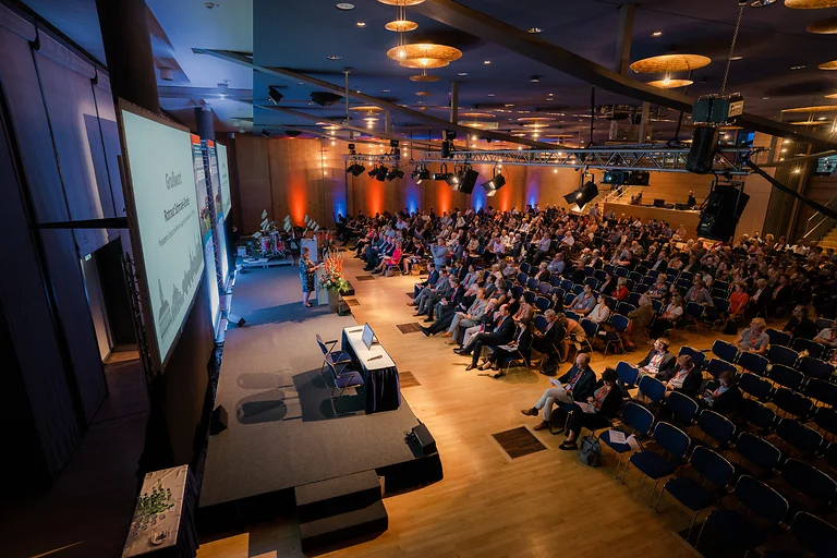 Blick von oben auf das Podium und die Sitzreihen voller Gäste, die begrüßt werden.