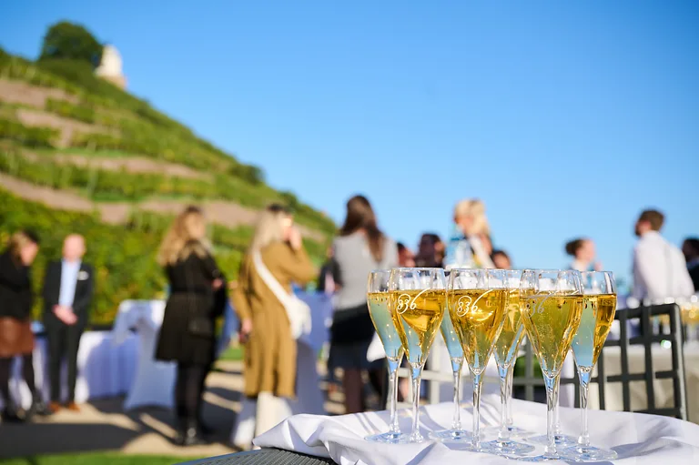 Sektempfang im Außenbereich von Schloss Wackerbarth bei blauem Himmel. Im Vordergrund rechts stehen gefüllte Sektgläser, im Hintergrund unterhalten sich Teilnehmende in entspannter Atmosphäre.