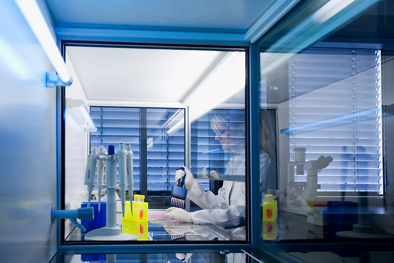 Laborantin mit Kittel und Handschuhen pipettiert Zellen in einer gläsernen Vorrichtung im Zellkulturraum des Bioinnovationszentrums Dresden.
