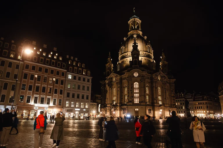 Neumarkt Dresden bei Nacht mit beleuchteter Frauenkirche im Hintergrund. Im Vordergrund laufen mehrere Personen über den Platz.