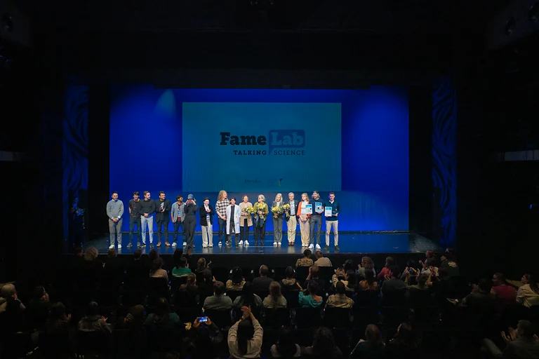 Alle Teilnehmerinnen, Teilnehmer und Juroren des FameLab 2025 stehen gemeinsam auf der Bühne im Kleinen Haus Dresden. Das Publikum applaudiert.