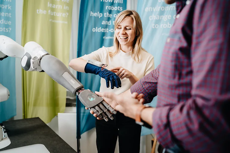 Eine lachende Frau testet den intelligenten Handschuh, im Vordergrund schüttelt eine Person einem Roboter die Hand.