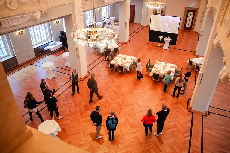 Blick von oben in den Löwensaal Dresden, Teilnehmende treffen ein, die Konferenztische sind vorbereitet und warten auf den Beginn der Veranstaltung.