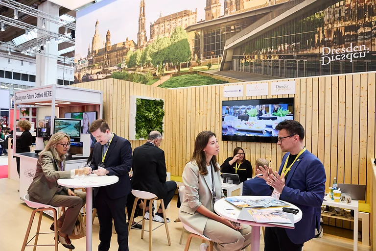 IMEX Frankfurt 2024, Mitarbeitende des Dresden Convention Teams im Gespräch mit Besuchenden an mehreren Tischen. Im Hintergrund ist der Barista Bot sichtbar, der für Aufmerksamkeit sorgt.