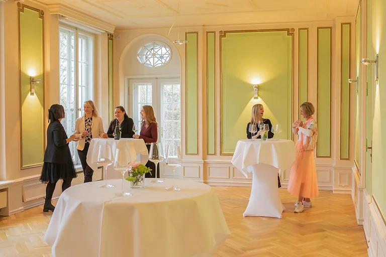 Gäste stehen an Stehtischen im Saal von Schloss Wackerbarth und genießen Gespräche bei einem Empfang.