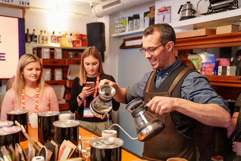 MCE 2023, Rahmenprogramm in der Kaffeerösterei. Ein Mann gießt Kaffee auf, während im Hintergrund Teilnehmende interessiert zuschauen.